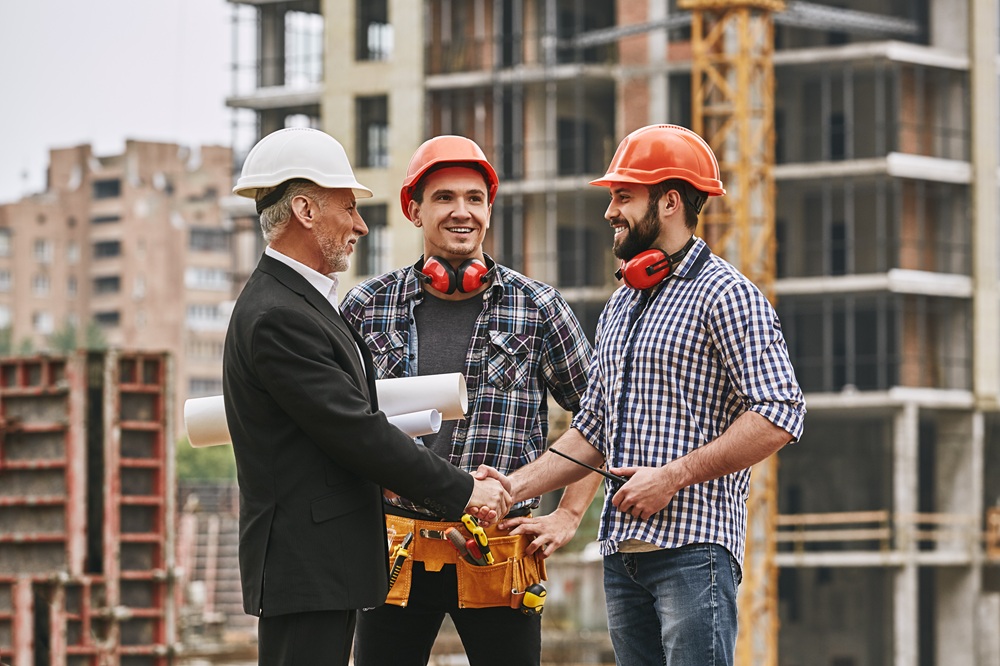 Good job! Cheerful elderly engineer in formal wear and white helmet holding engineering drawings and shaking hands of two happy young builders while standing at construction site.